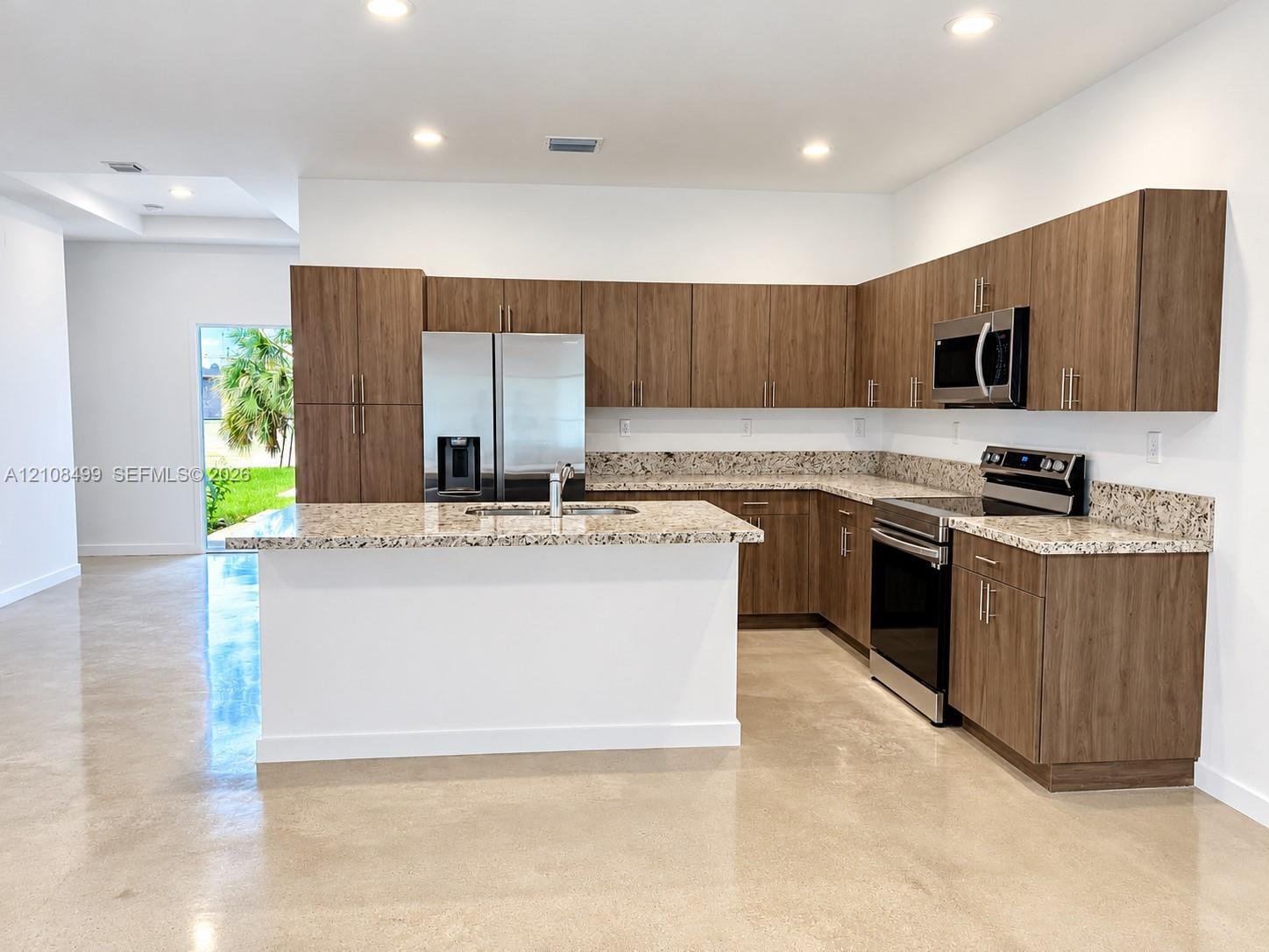 Kitchen featuring wood-finish cabinetry, stone-surface countertops, central island with an integrated sink, stainless steel appliances, and polished concrete flooring
