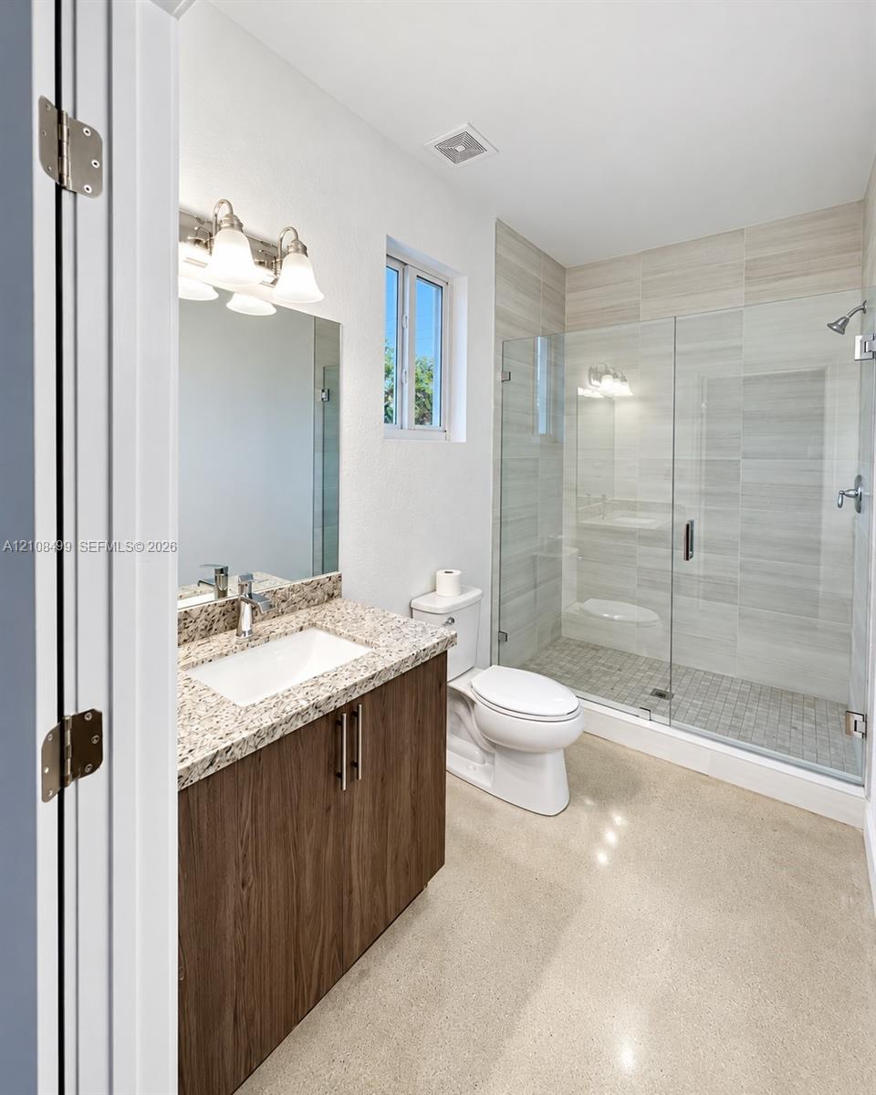 Bathroom featuring a wood-finish vanity with a stone countertop, an integrated rectangular sink, a glass-enclosed shower with tile surround, and polished concrete flooring