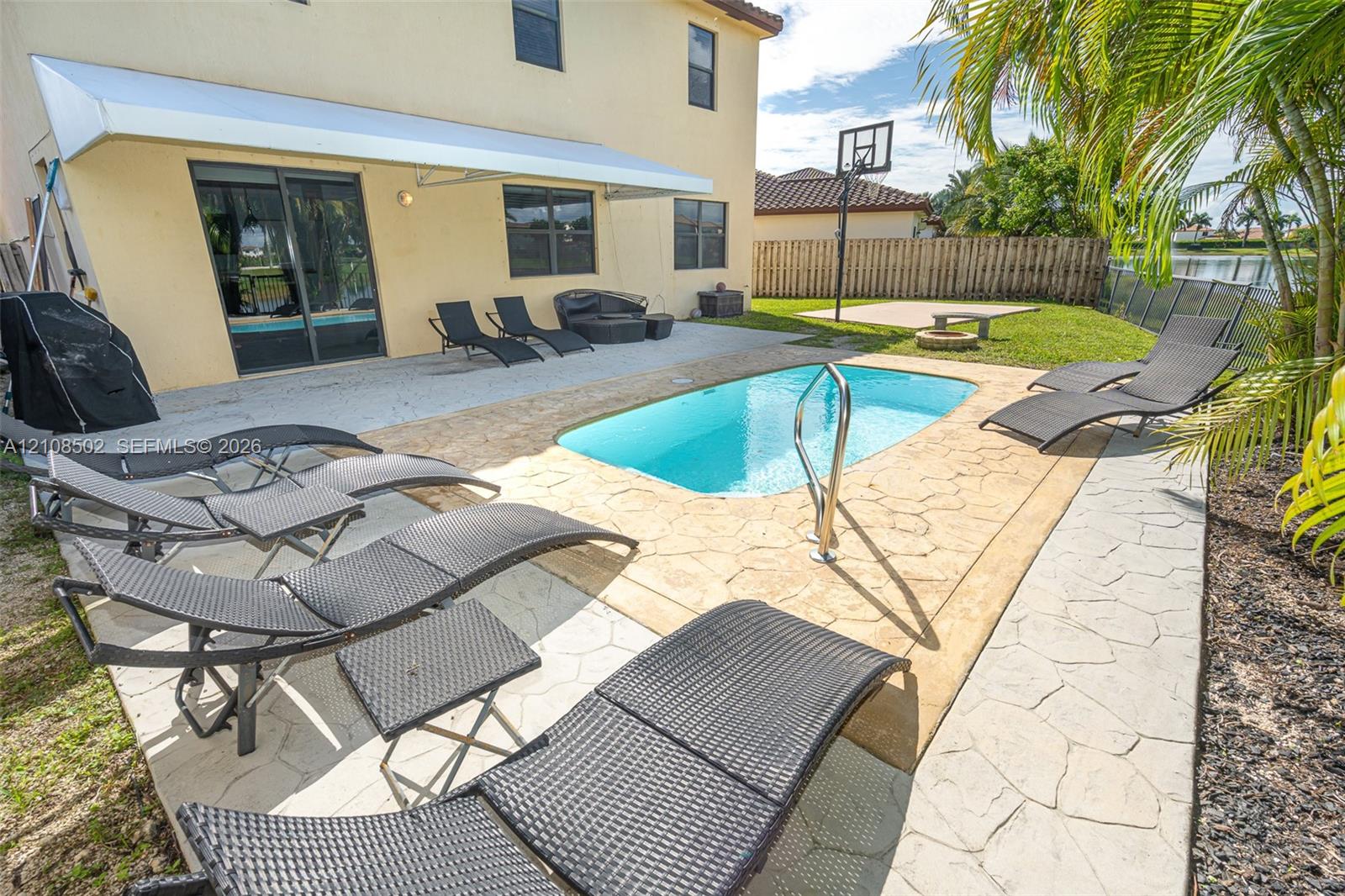 Backyard featuring a concrete pool with a metal handrail, a stamped concrete patio, a retractable awning, and a built-in basketball hoop
