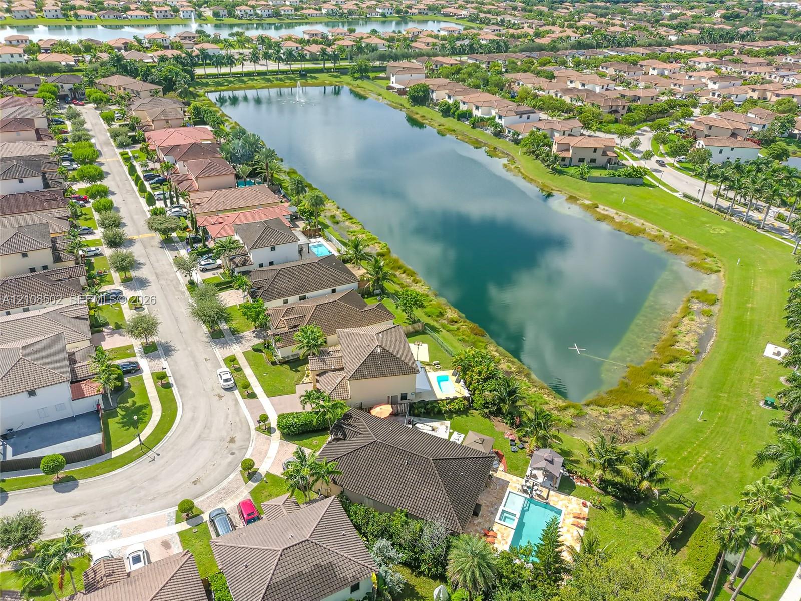 Aerial perspective of a residential property featuring an inground swimming pool, stone-paver pool deck, mature landscaping, and placement along a waterfront pond