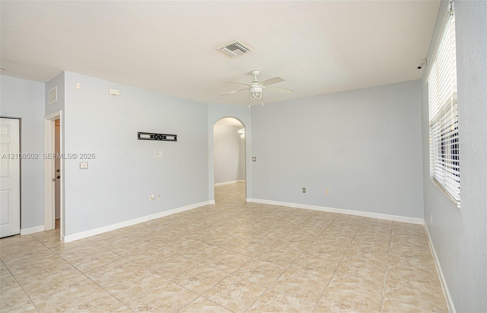 Neutral-toned interior space featuring light-colored tile flooring, an arched doorway, a ceiling fan with integrated lighting, and horizontal window blinds