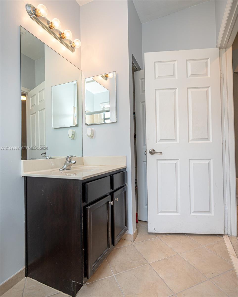 Dark-toned cabinetry vanity with integrated countertop, single-handle faucet, large wall-mounted mirror, four-bulb light fixture, and tile flooring
