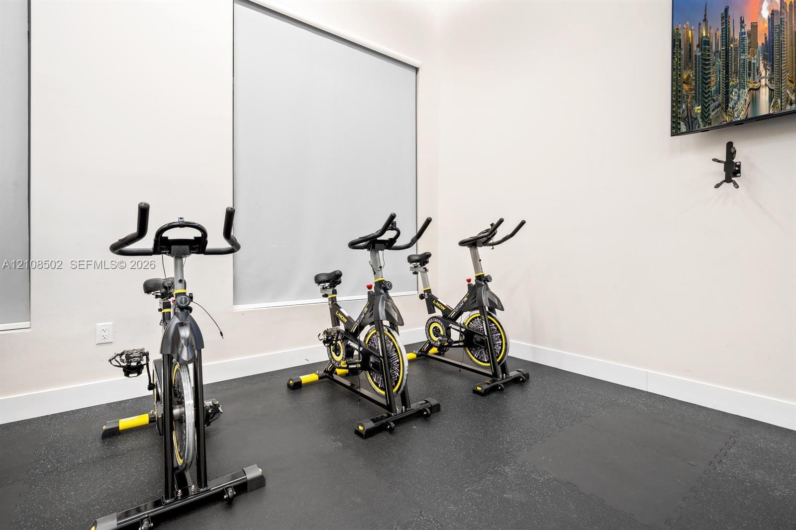 Dedicated exercise space featuring durable rubberized flooring, large windows with neutral roller shades, and wall-mounted television hardware