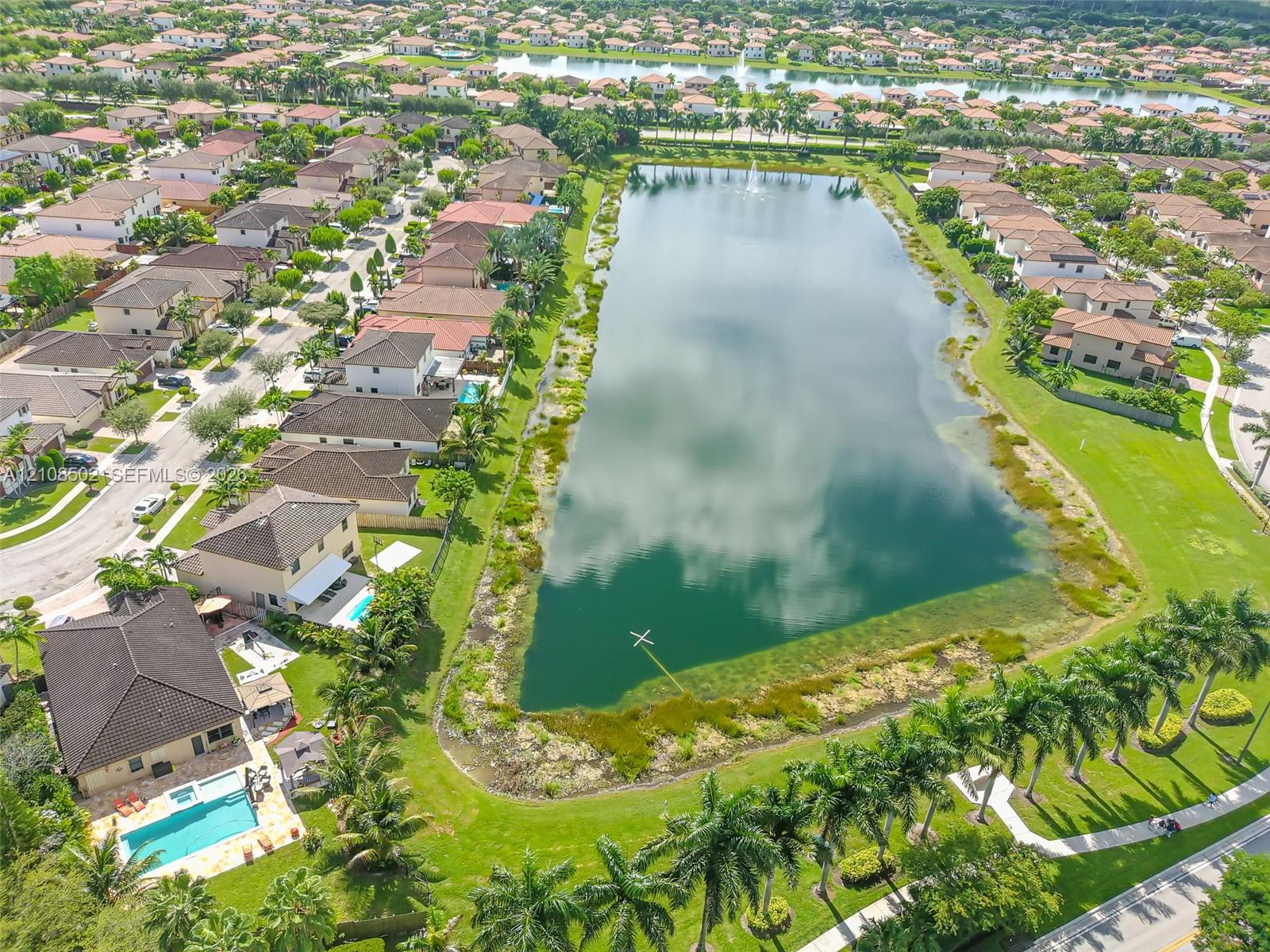 Aerial perspective featuring a multi-story residence with a paved pool deck, rectangular swimming pool, manicured lawn, and proximity to a large central lake