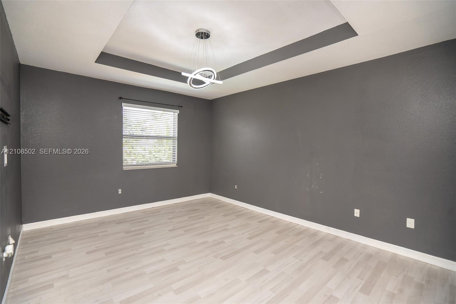 Tray ceiling featuring a contemporary circular chandelier, light-toned wood-finish flooring, and single-pane window with installed blinds