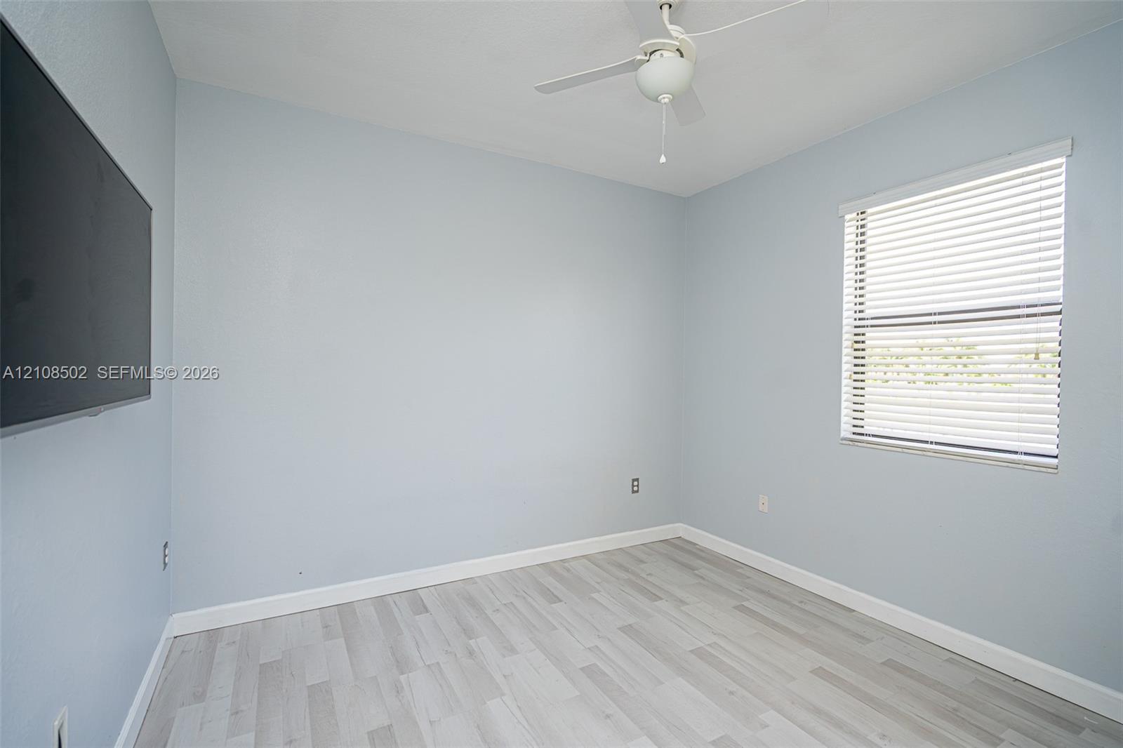 Neutral wall-toned room featuring wood-finish flooring, a white ceiling fan, and a window with horizontal blinds