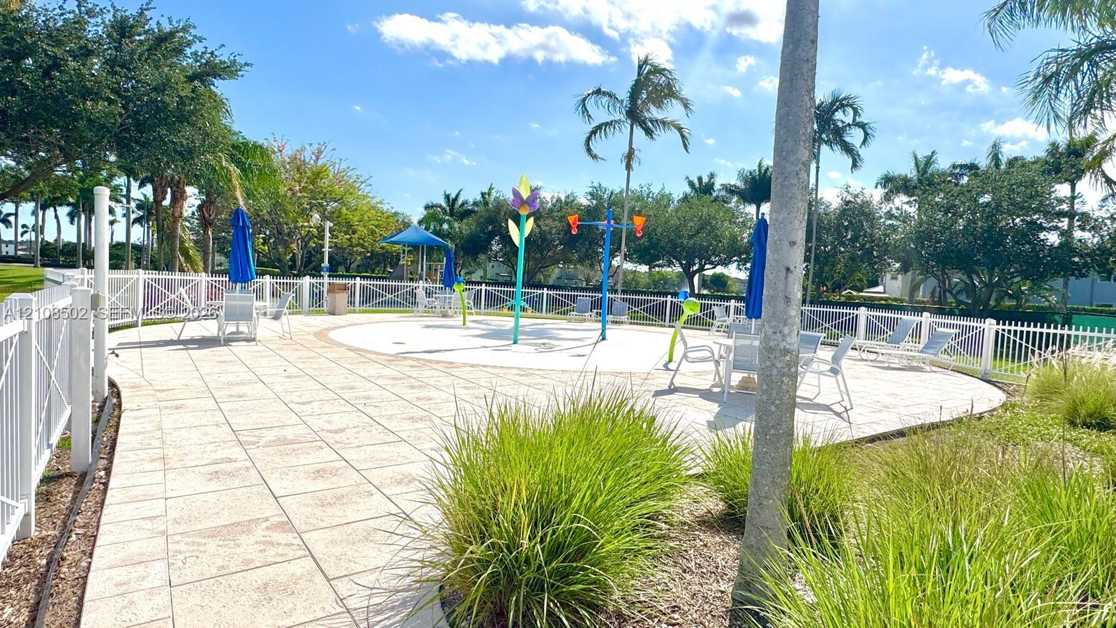 Outdoor splash pad featuring water features, paved patio decking, white perimeter fencing, and mature palm trees