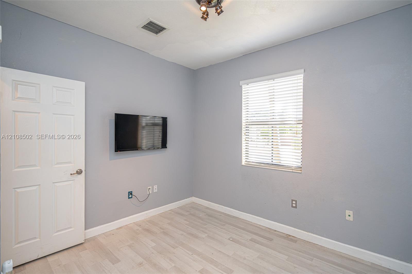 Interior room featuring wood-finish flooring, a multi-panel door with a metallic handle, a window equipped with horizontal blinds, and a mounted adjustable television bracket