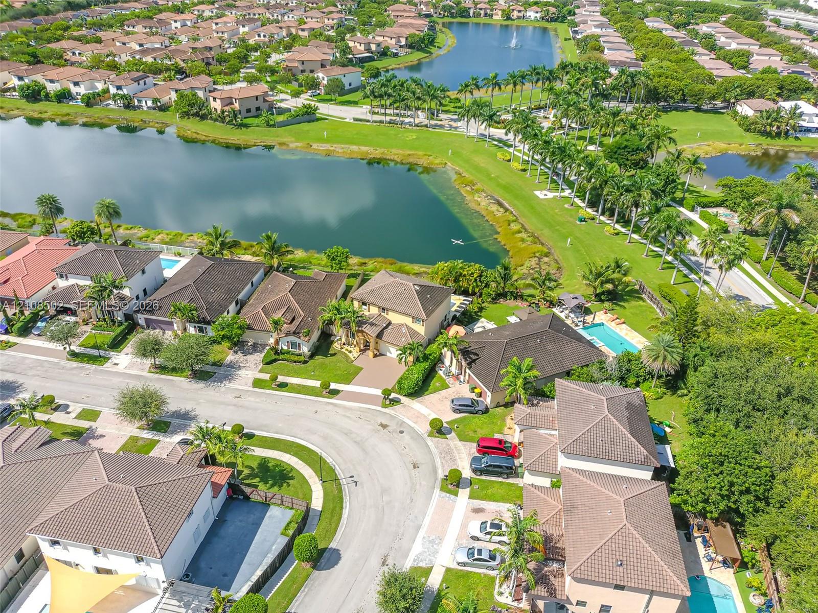 Aerial perspective featuring a multi-parcel residential community with winding paved streets, manicured green space, mature landscaping, and a central lake