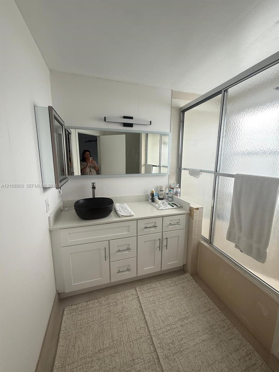 White cabinetry vanity featuring a solid surface countertop, matte black vessel sink, wall-mounted rectangular mirror, and a sliding glass shower door White cabinetry vanity featuring a solid surface countertop, matte black vessel sink, wall-mounted rectangular mirror, and a sliding glass shower door