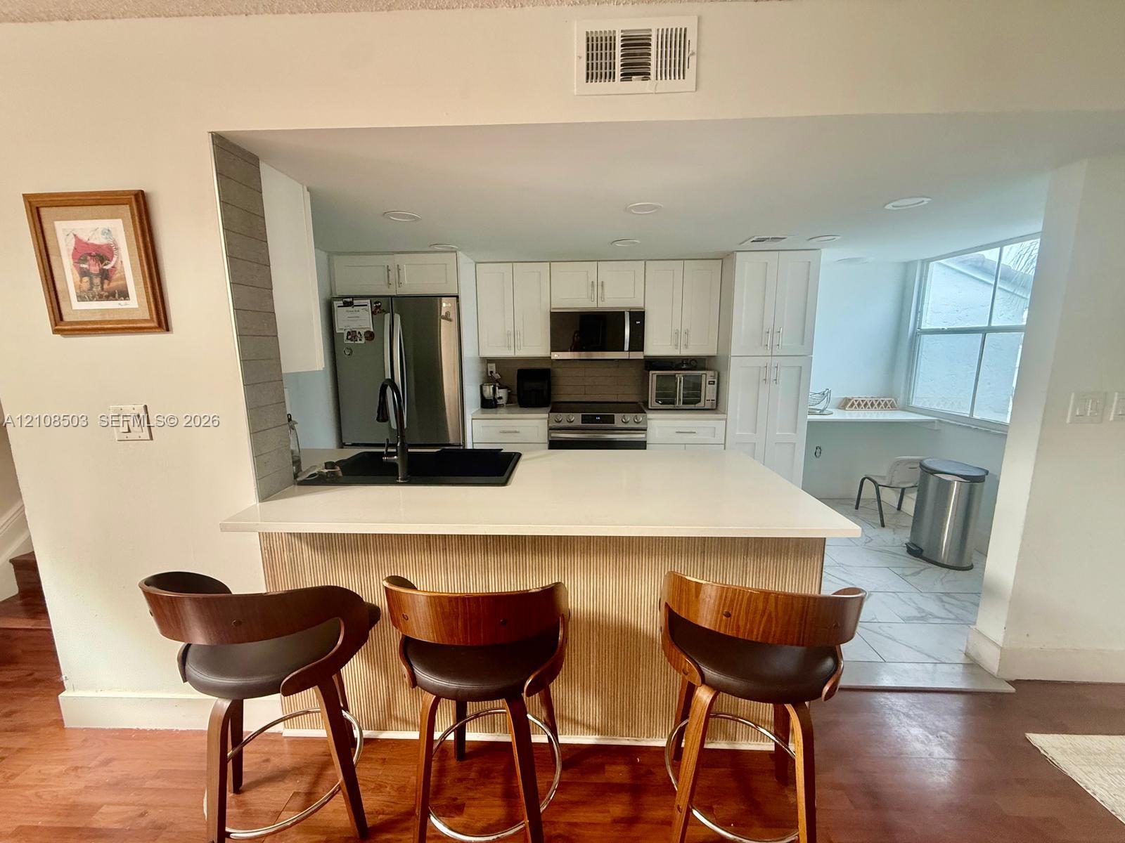 Kitchen featuring white cabinetry, solid-surface countertops, integrated stainless steel appliances, a black undermount sink with a matte black faucet, and wood-finish cabinetry detailing Kitchen featuring white cabinetry, solid-surface countertops, integrated stainless steel appliances, a black undermount sink with a matte black faucet, and wood-finish cabinetry detailing