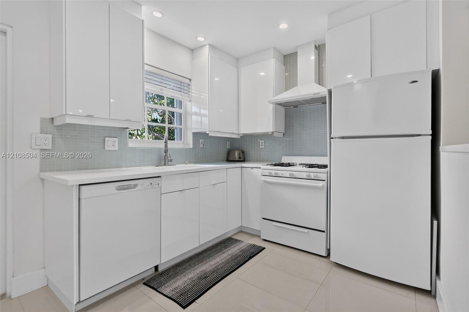 Kitchen featuring flat-panel white cabinetry, light-toned tile flooring, light blue glass tile backsplash, white countertops, and a suite of white appliances