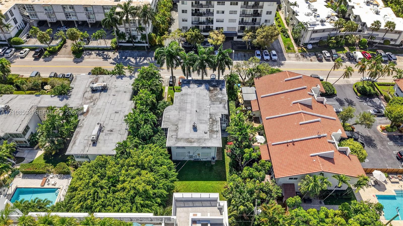 Aerial view of urban property featuring a flat roof structure, mature landscaping, palm trees, and ground-level grass coverage