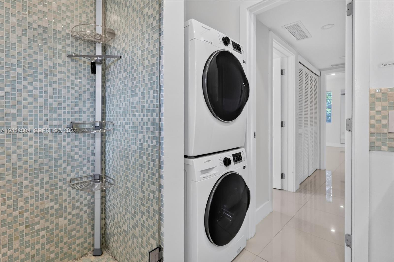 Laundry area featuring stacked front-load washer and dryer, tiled wall with integrated shelving, and polished neutral-tone flooring