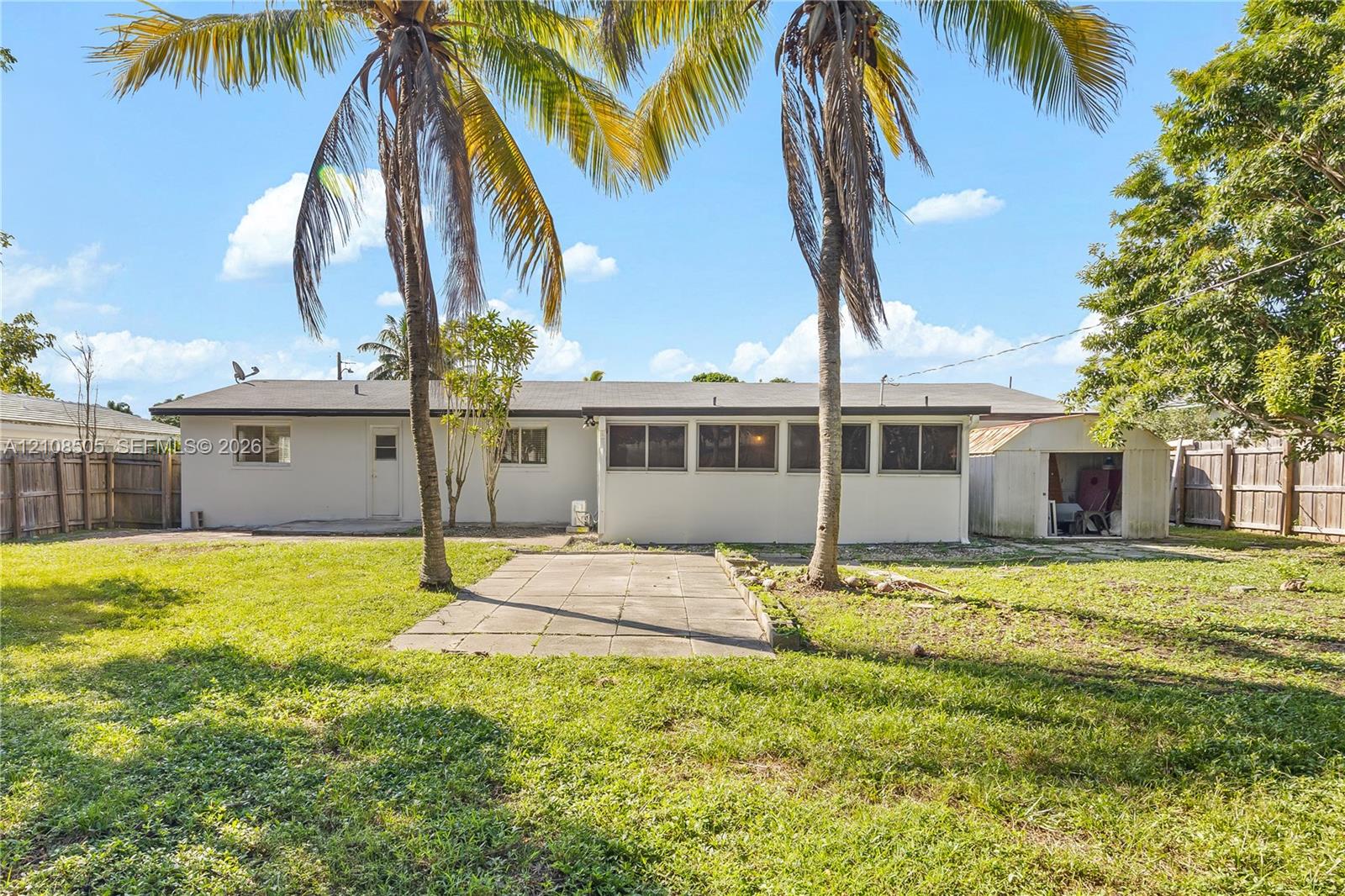 Spacious backyard featuring a grass lawn, paved patio, mature palm trees, detached storage structure, and wood privacy fencing Spacious backyard featuring a grass lawn, paved patio, mature palm trees, detached storage structure, and wood privacy fencing