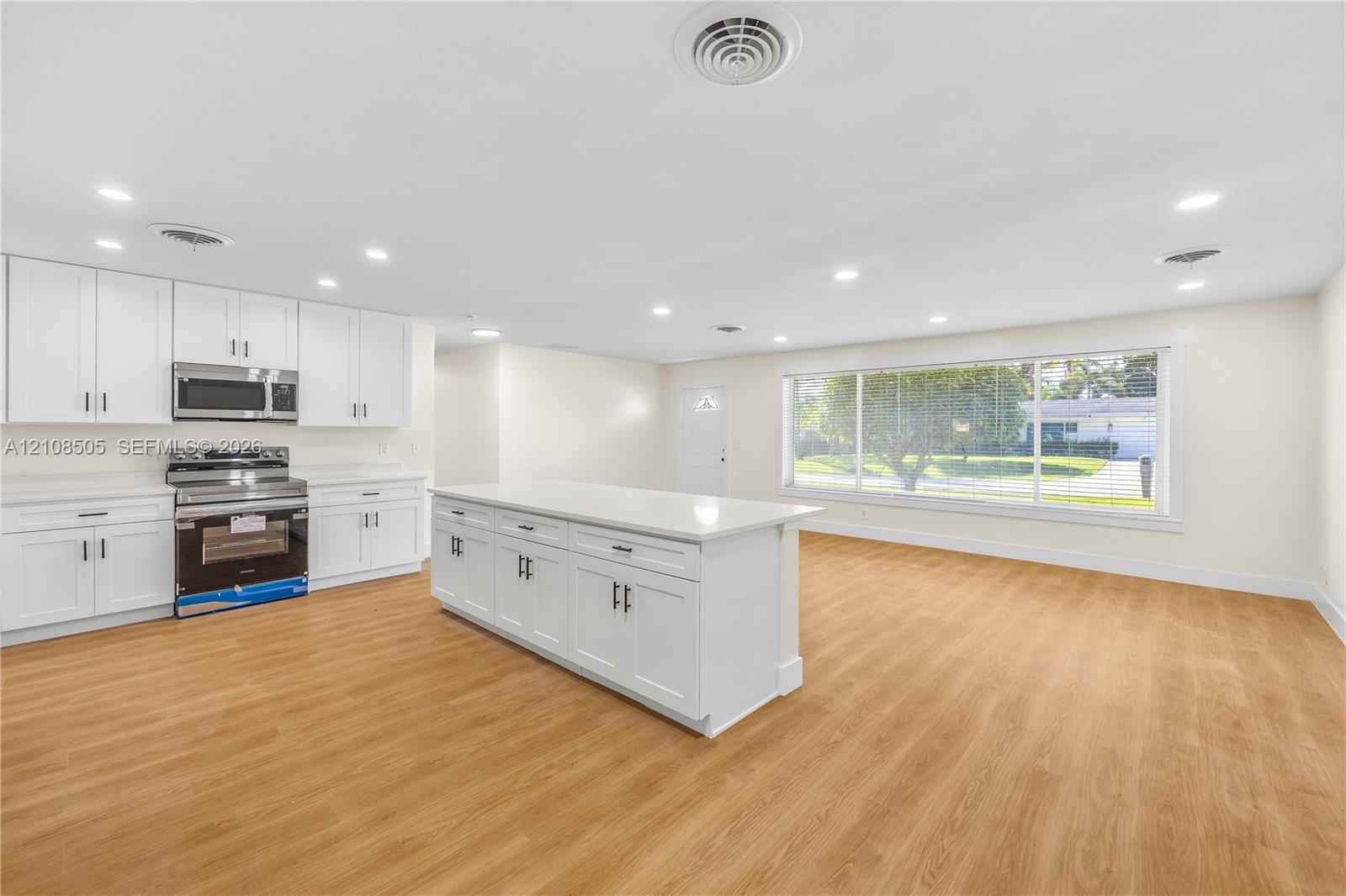 Open-concept kitchen featuring white shaker-style cabinetry, a central island with a solid-surface countertop, stainless steel appliances, recessed lighting, and wood-finish flooring Open-concept kitchen featuring white shaker-style cabinetry, a central island with a solid-surface countertop, stainless steel appliances, recessed lighting, and wood-finish flooring