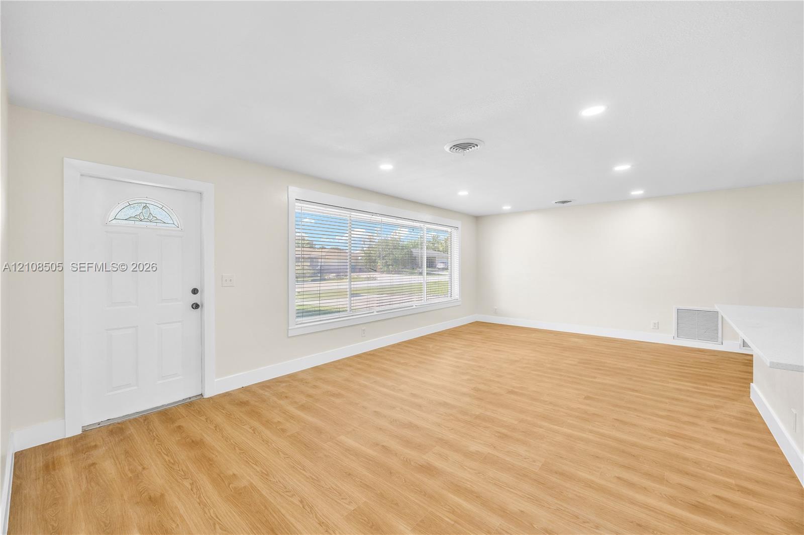 Open-concept area featuring wood-finish flooring, recessed lighting, a wide window with horizontal blinds, and a white entry door with a decorative glass insert Open-concept area featuring wood-finish flooring, recessed lighting, a wide window with horizontal blinds, and a white entry door with a decorative glass insert