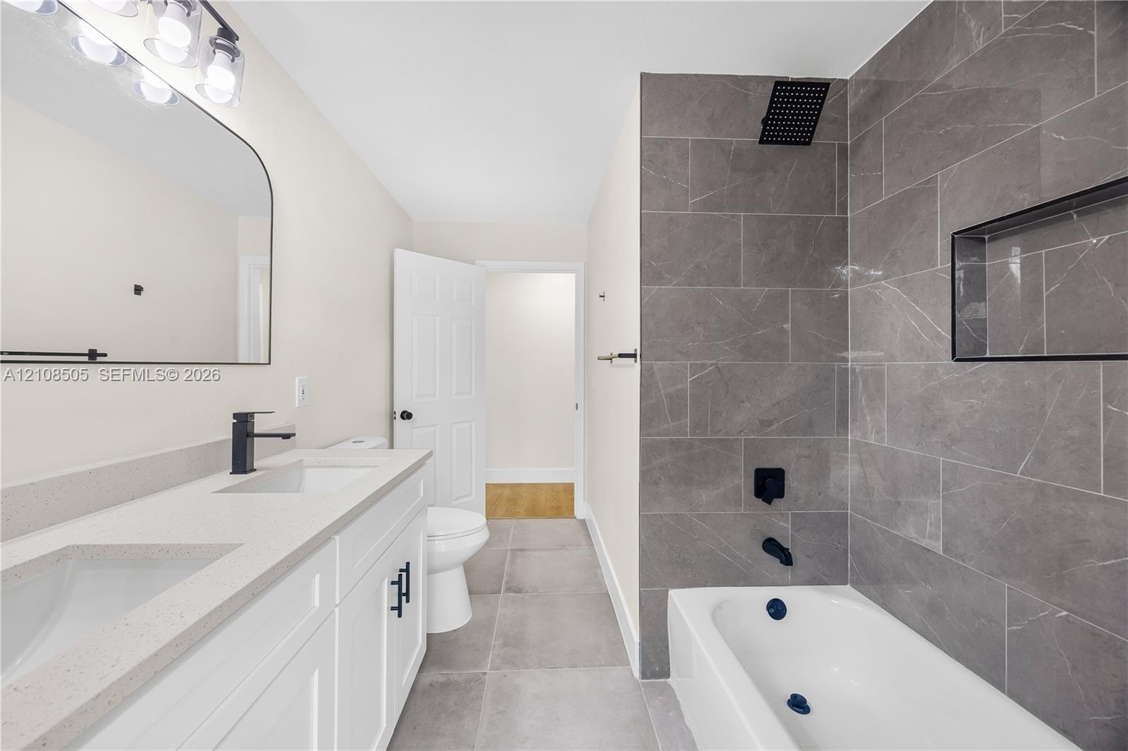 Double vanity with a stone-finish countertop, matte black plumbing fixtures, arched wall mirror, and a bathtub with full-height stone-finish tile surround and built-in recessed niche Double vanity with a stone-finish countertop, matte black plumbing fixtures, arched wall mirror, and a bathtub with full-height stone-finish tile surround and built-in recessed niche