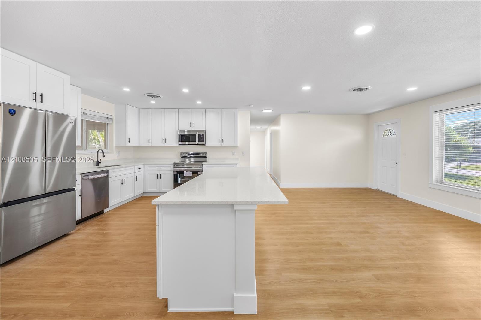 Kitchen featuring white cabinetry, an expansive central island with a solid-surface countertop, stainless steel appliances, and wood-finish flooring with recessed lighting Kitchen featuring white cabinetry, an expansive central island with a solid-surface countertop, stainless steel appliances, and wood-finish flooring with recessed lighting