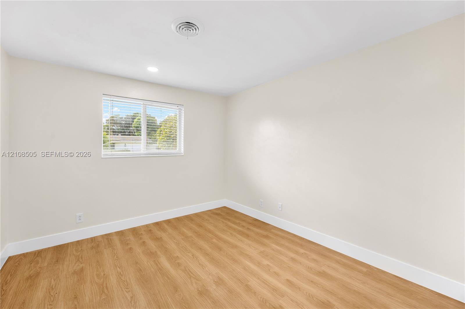 Neutral-toned interior space featuring wood-finish flooring, crisp white wall paint, and a single window with horizontal blinds