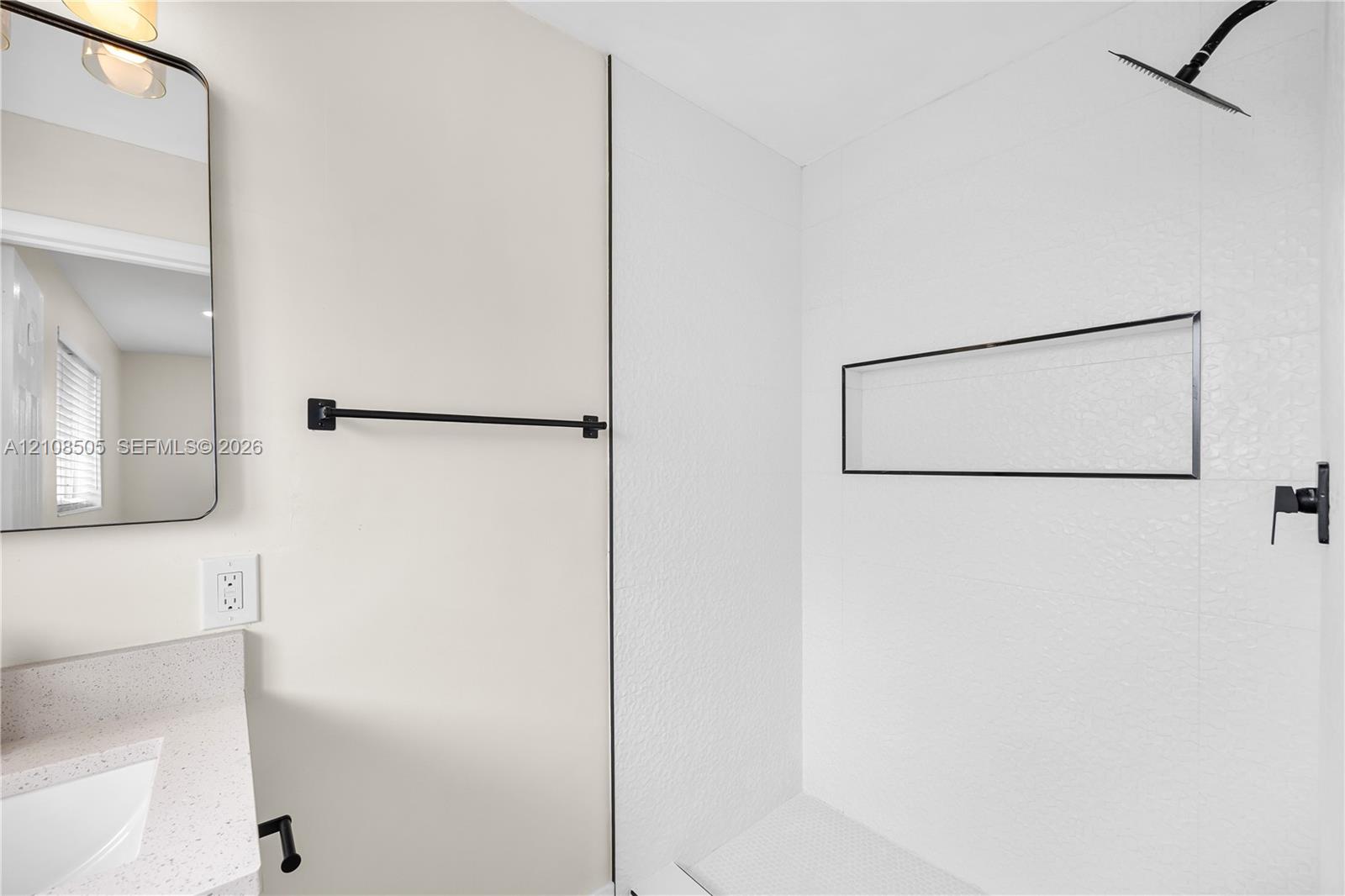 Bathroom featuring a walk-in shower with textured tile surround and built-in recessed shelf, matte black wall-mounted shower head and fixtures, stone-finish countertop with integrated sink, and an arched wall-mounted mirror