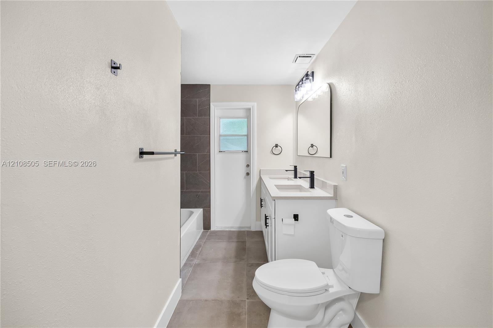 Dual-vanity cabinetry with rectangular integrated sinks, matte black fixtures, neutral large-format floor tiling, and a tub enclosure with full-height tile surround