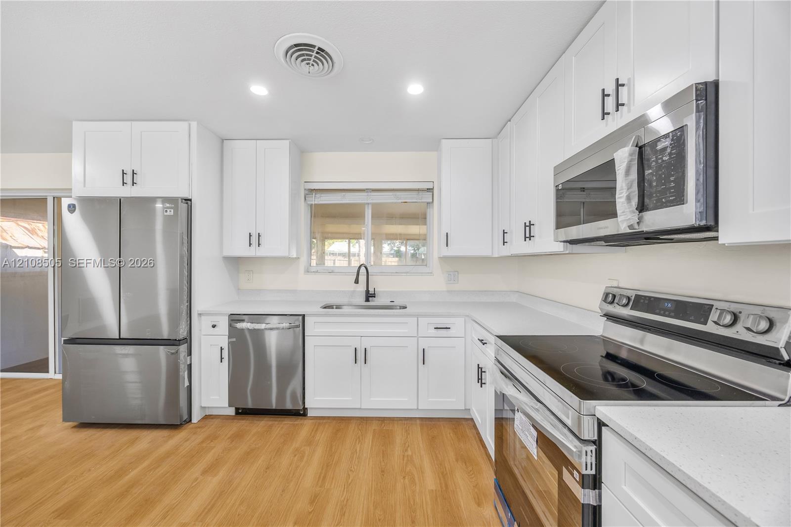 Kitchen featuring white shaker-style cabinetry, solid-surface countertops, stainless steel appliances, matte black hardware, and wood-finish flooring Kitchen featuring white shaker-style cabinetry, solid-surface countertops, stainless steel appliances, matte black hardware, and wood-finish flooring