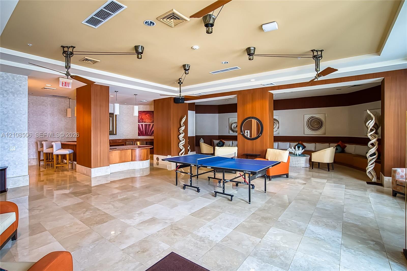 Large common area featuring natural stone tile flooring, wood-paneled columns, built-in bar with stone countertop, recessed lighting, and dual ceiling-mounted fans