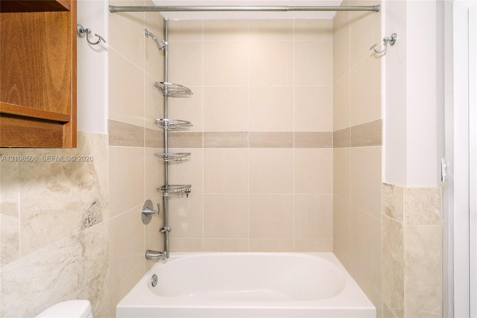 Shower area featuring floor-to-ceiling neutral ceramic tiling, an integrated stone-finish accent band, a built-in recessed tub, chrome plumbing fixtures, and a wall-mounted metal shelving unit