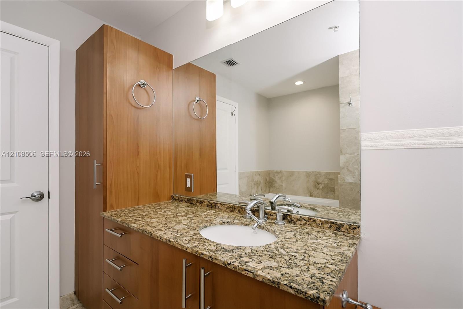 Wood-finish cabinetry with integrated tall storage, stone-surfaced vanity top, undermount sink, and large wall-mounted mirror