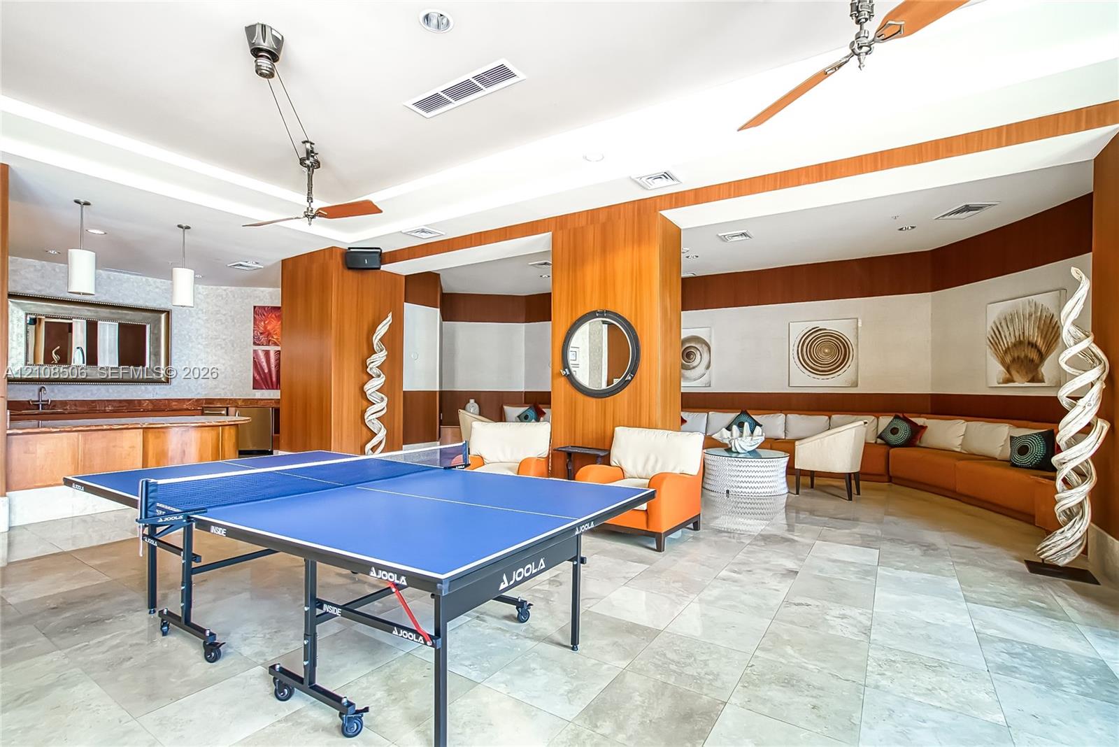 Large recreation room featuring stone tile flooring, built-in wet bar with stone countertop, wood-panelled accent walls, integrated ceiling fans, and recessed lighting