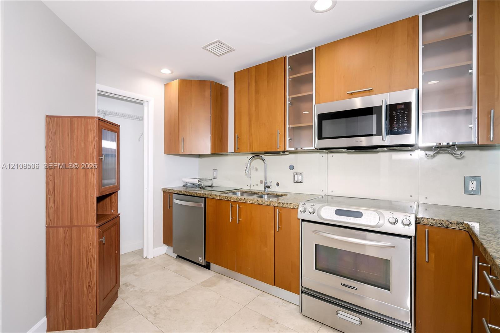 Kitchen featuring natural wood-finish cabinetry, stone-surface countertops, stainless steel appliances, glass-panel cabinet inserts, and tile flooring