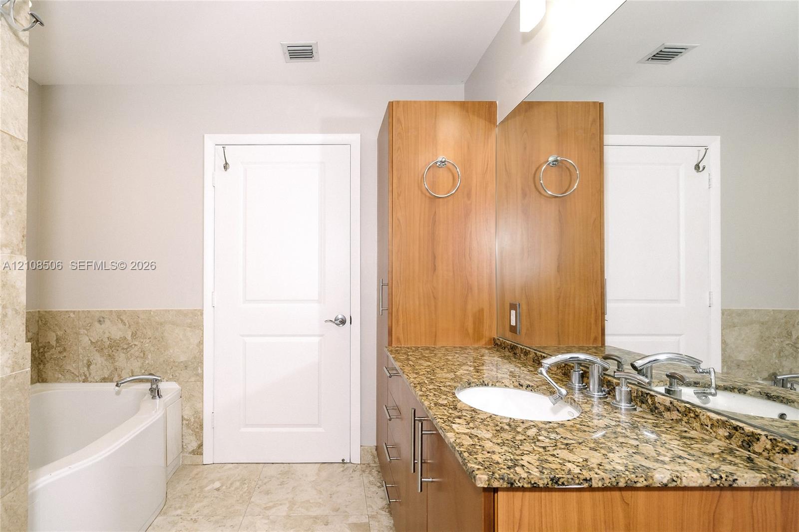 Bathroom featuring stone-surface vanity with dual integrated sinks, floor-to-ceiling wood cabinetry, large wall-mounted mirror, and a soaking tub with stone tile surround