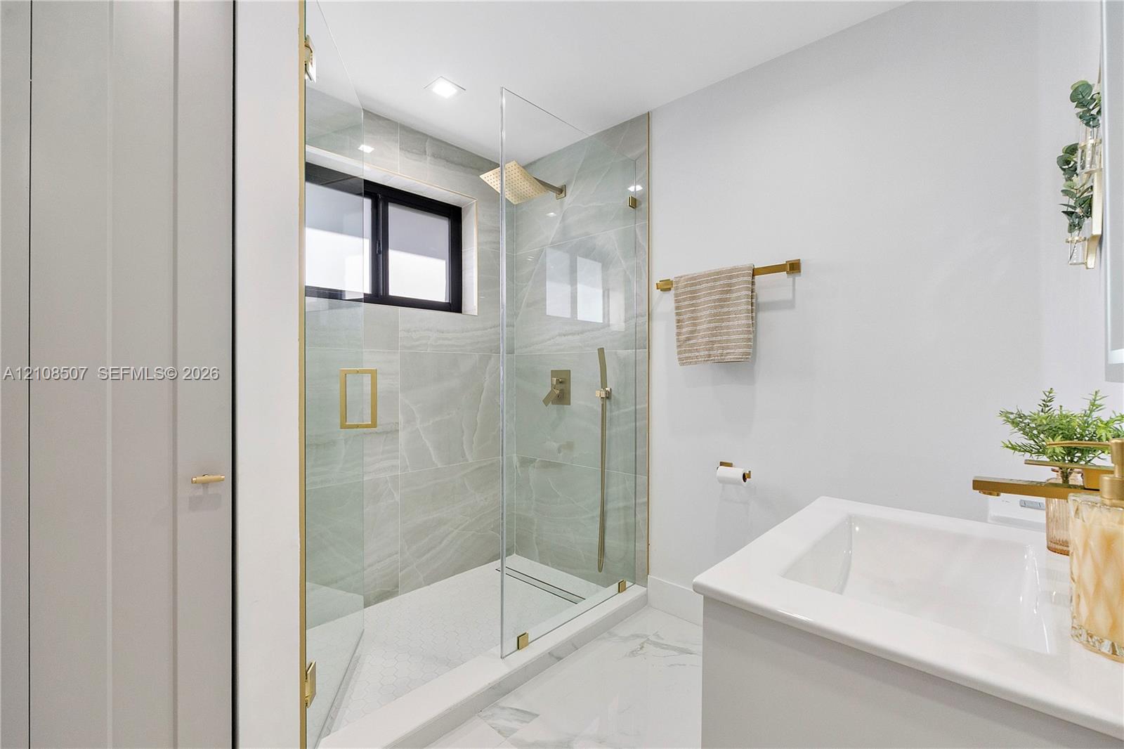 Walk-in shower featuring large-format tile surround, glass enclosure, brass-finish fixtures, and a recessed window