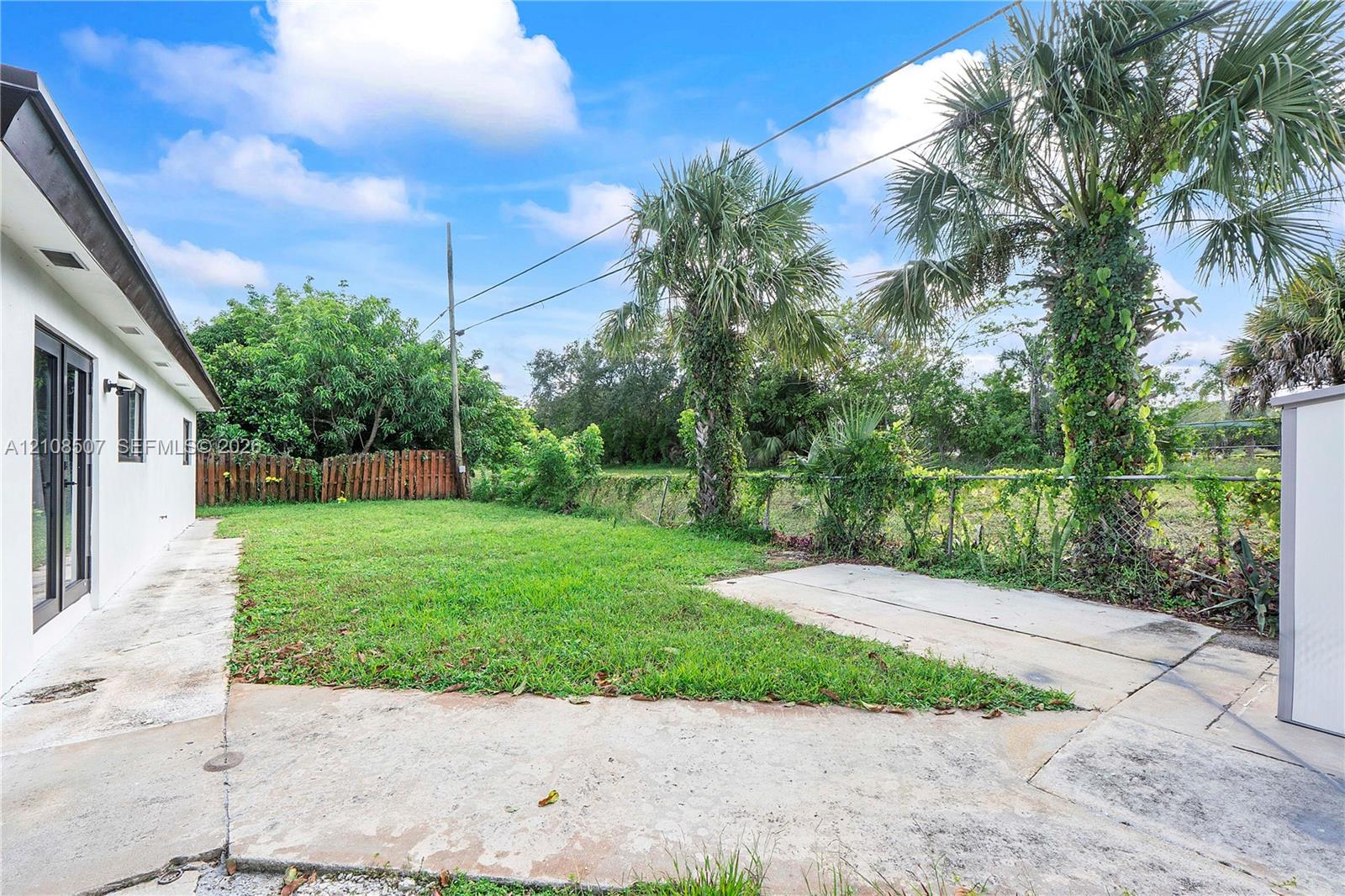 Rear exterior featuring a concrete patio, grassy lawn, mature palm trees, and perimeter fencing