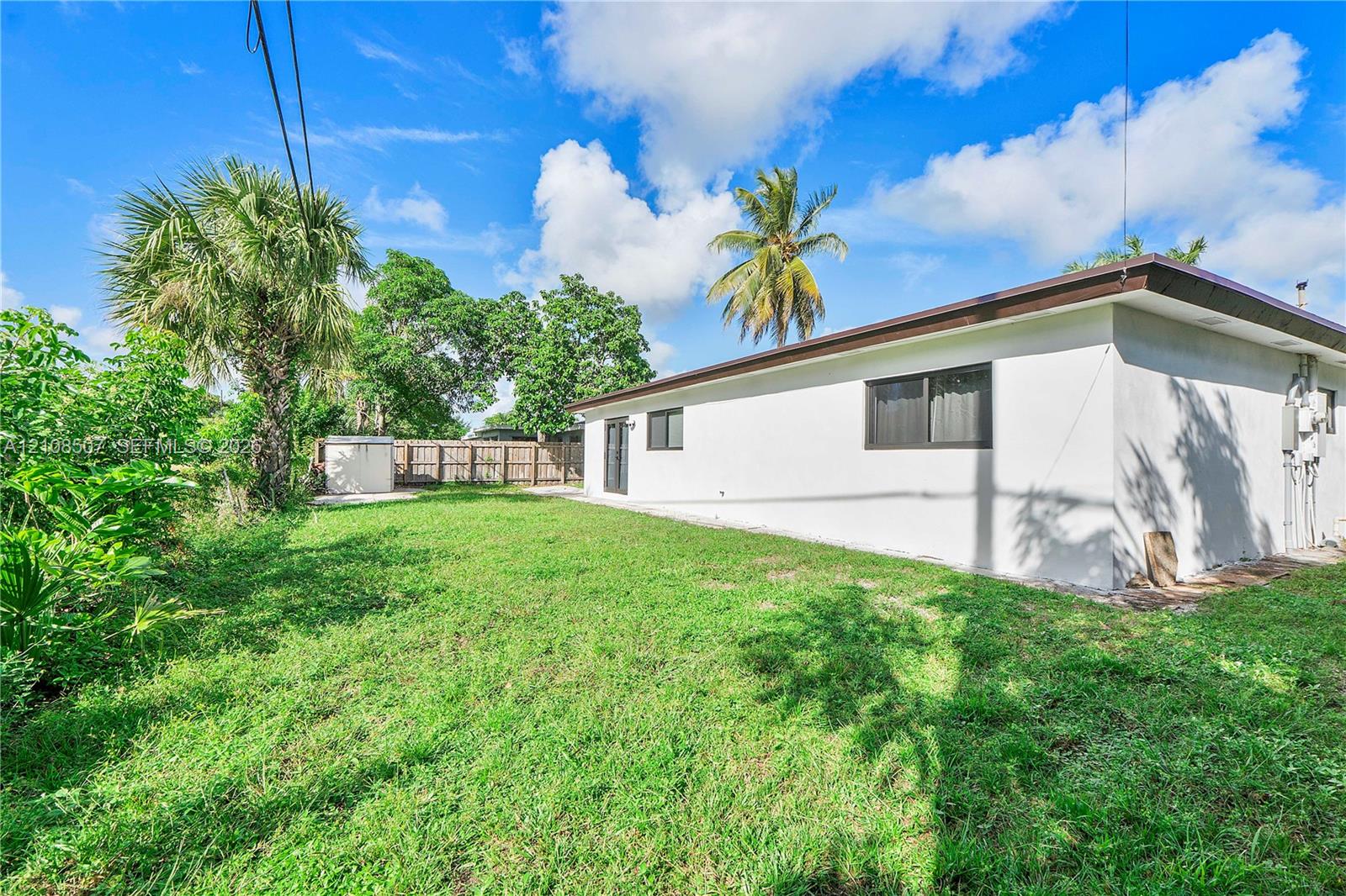 Exterior of a single-story residence featuring a smooth stucco facade, dark-framed windows, and a private yard enclosed by wood fencing with mature palm trees