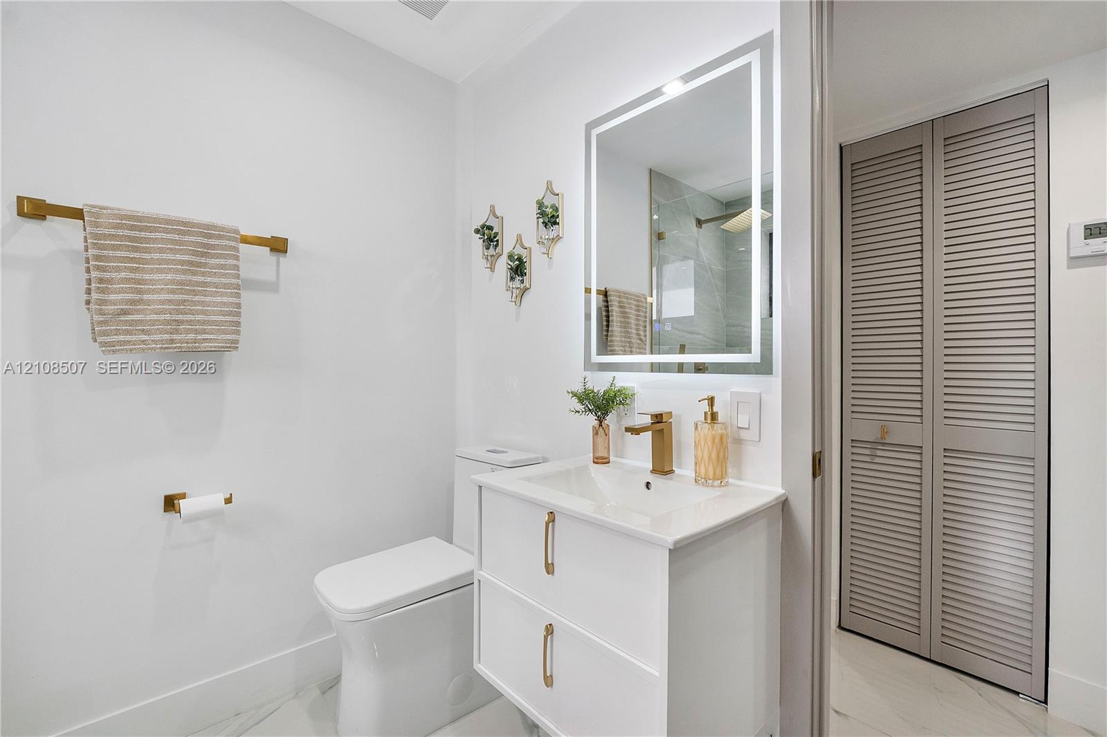 White vanity with a rectangular sink, quartz-style countertop, brass-finish faucet and hardware, LED-backlit mirror, and louvered-panel closet doors