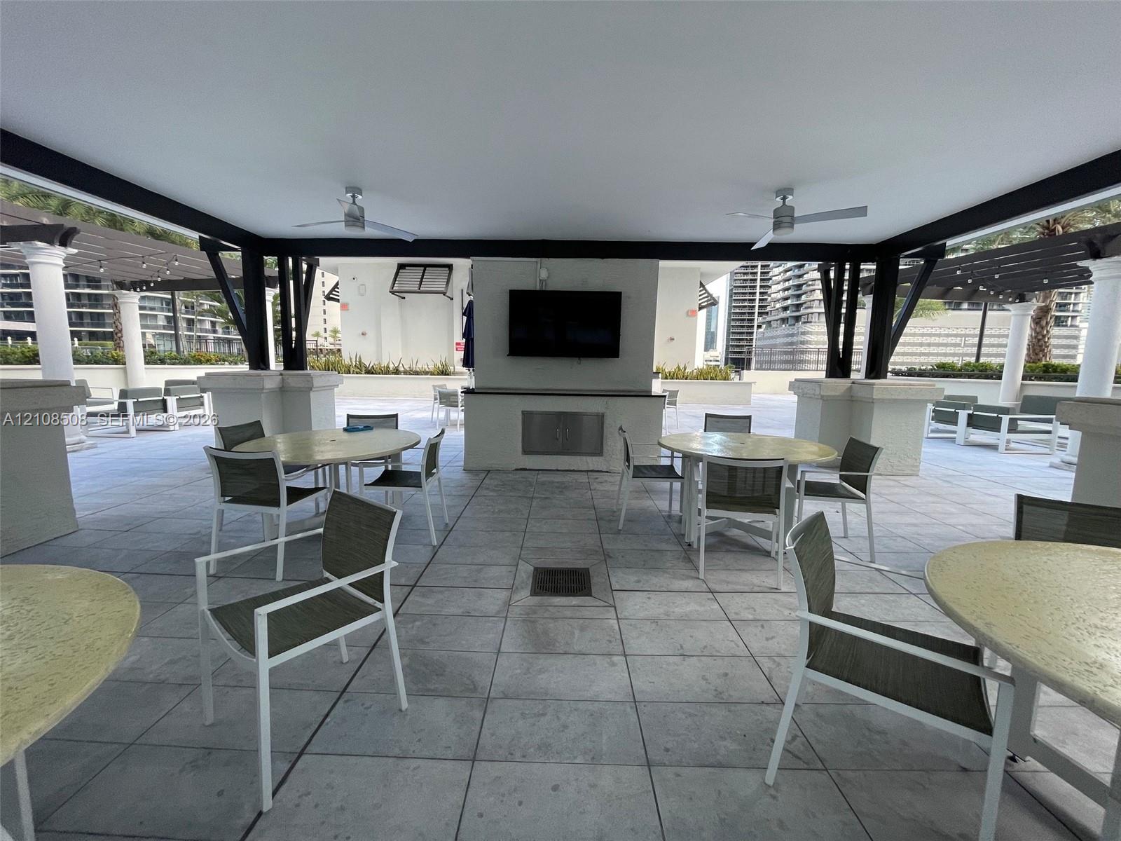 Covered outdoor terrace featuring a tiled floor, dual ceiling fans, built-in structural columns, and a central wall mount for electronics