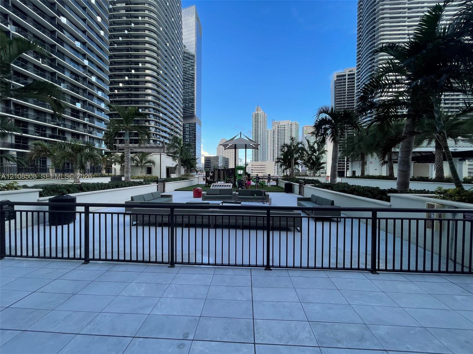 Elevated terrace featuring light-toned stone tile flooring, perimeter metal railings, integrated landscaping, and palm trees set against an urban skyline backdrop