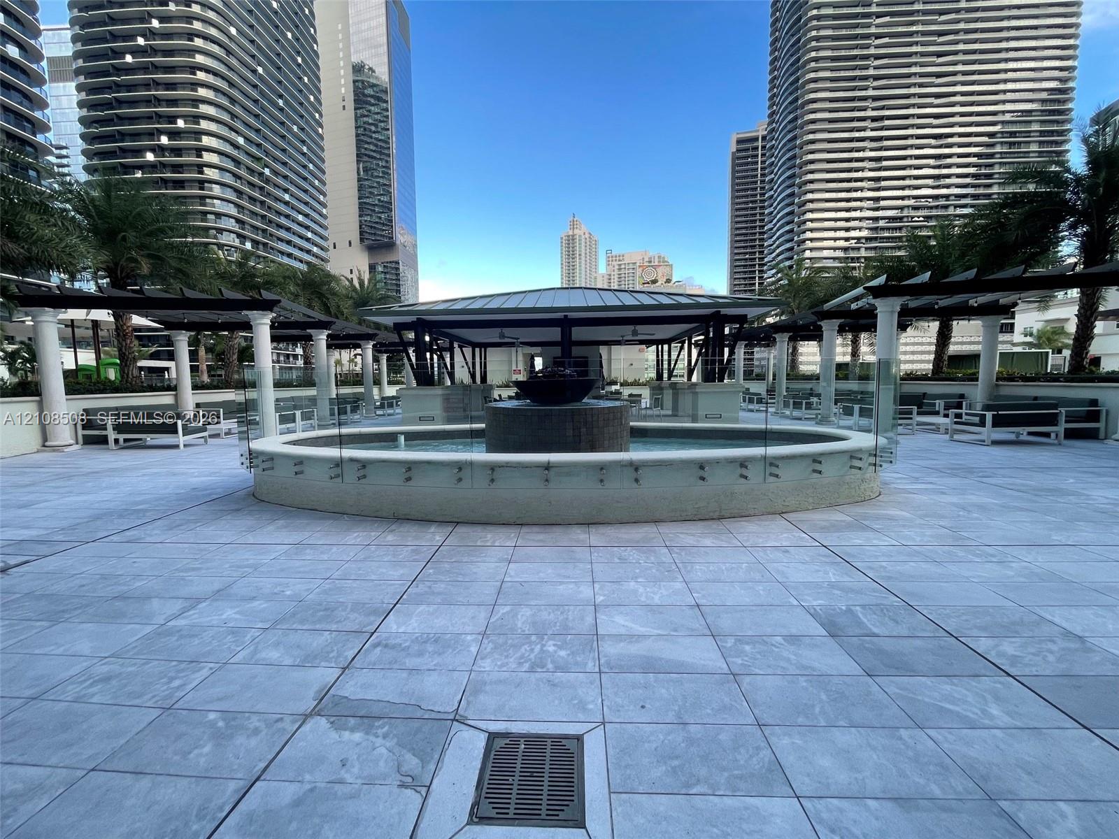 Outdoor terrace featuring a central stone-clad fountain, stone tile flooring, covered pergolas with structural columns, and glass safety railings