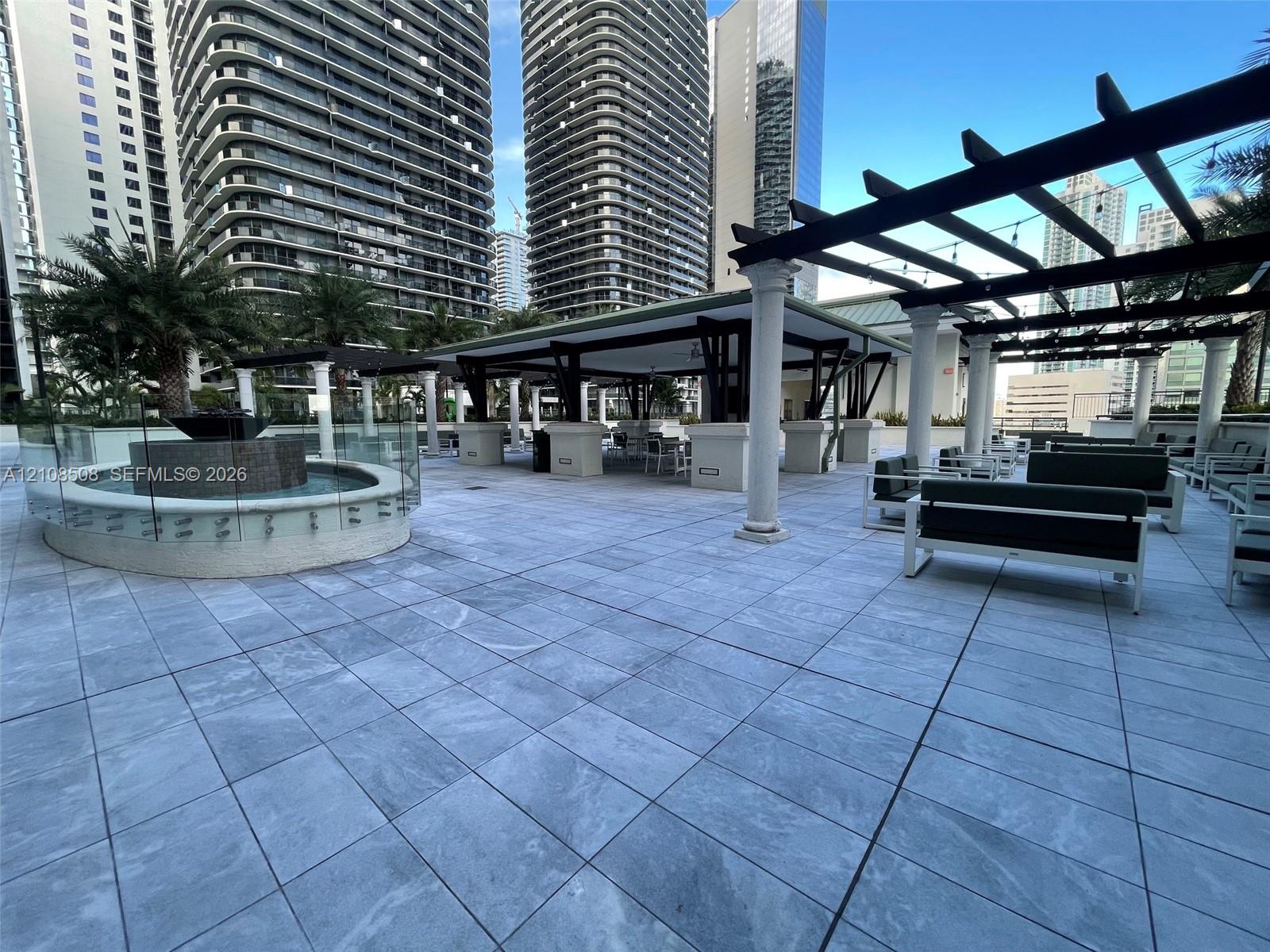 Outdoor terrace featuring large-format stone-look tiling, a circular stone-clad water feature with glass surround, and a covered pergola supported by classic columns