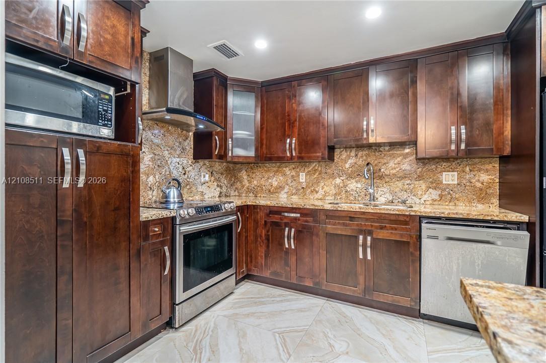 Kitchen featuring dark wood cabinetry, stone countertops, stone backsplash, stainless steel appliances, and large-format tile flooring Kitchen featuring dark wood cabinetry, stone countertops, stone backsplash, stainless steel appliances, and large-format tile flooring