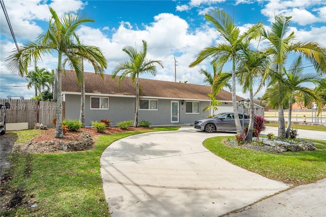 Single-story exterior featuring a concrete driveway, established palm trees, landscaped garden beds, and a wood fence Single-story exterior featuring a concrete driveway, established palm trees, landscaped garden beds, and a wood fence