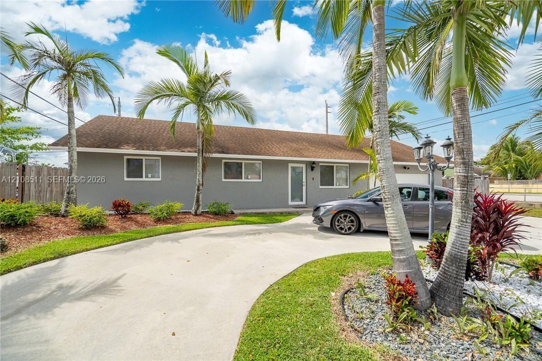 Single-story exterior featuring a gray stucco facade, arched concrete driveway, multi-pane windows, and landscaped grounds with mature palm trees Single-story exterior featuring a gray stucco facade, arched concrete driveway, multi-pane windows, and landscaped grounds with mature palm trees