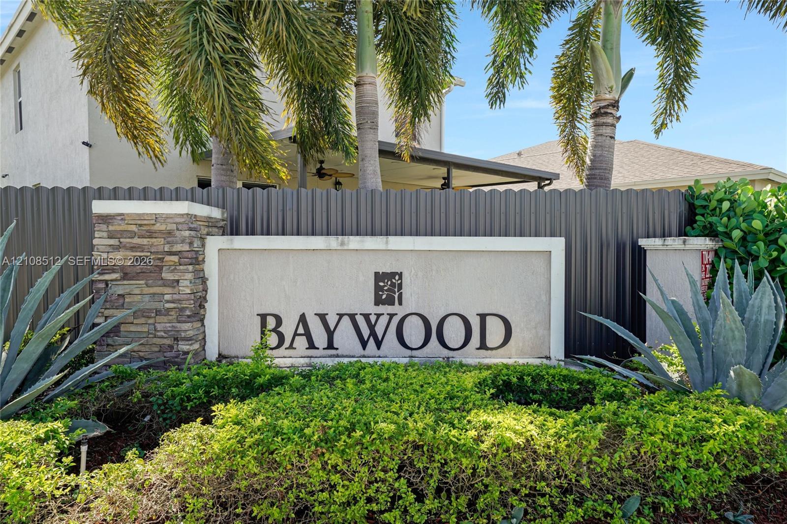Baywood community entrance featuring a masonry monument sign, stacked stone accents, mature palm trees, and professional landscaping