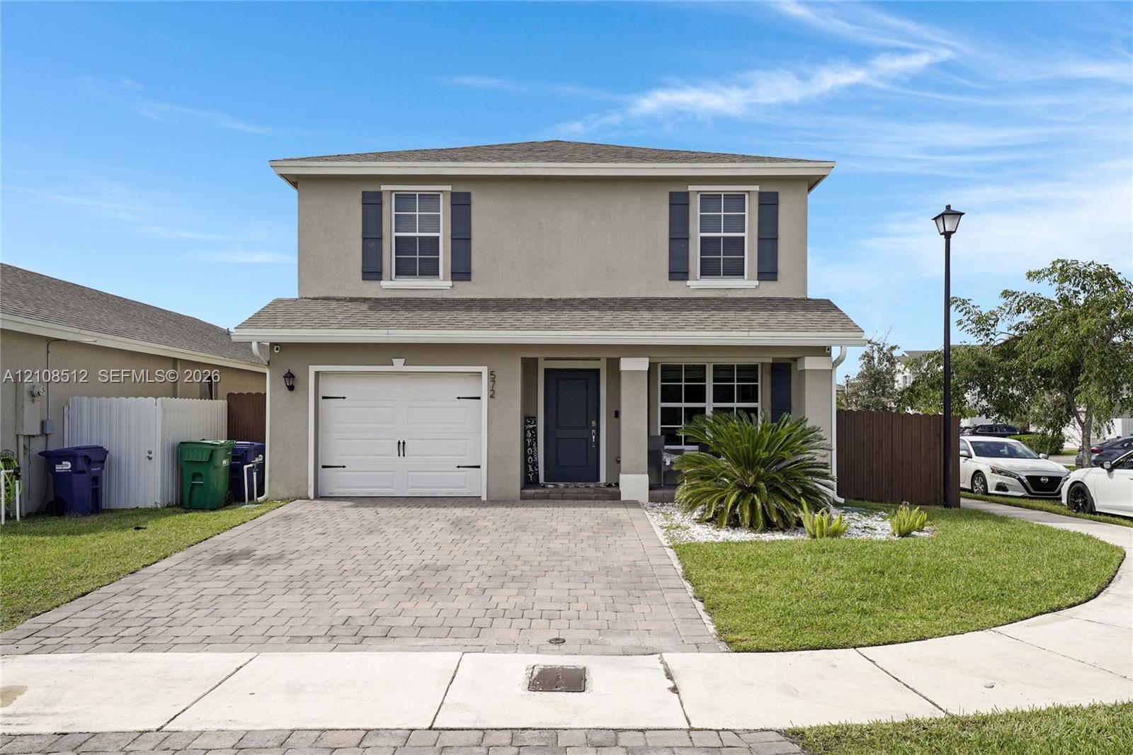 Two-story exterior featuring a neutral stucco facade, grey decorative shutters, a single-car garage, and a paved stone driveway