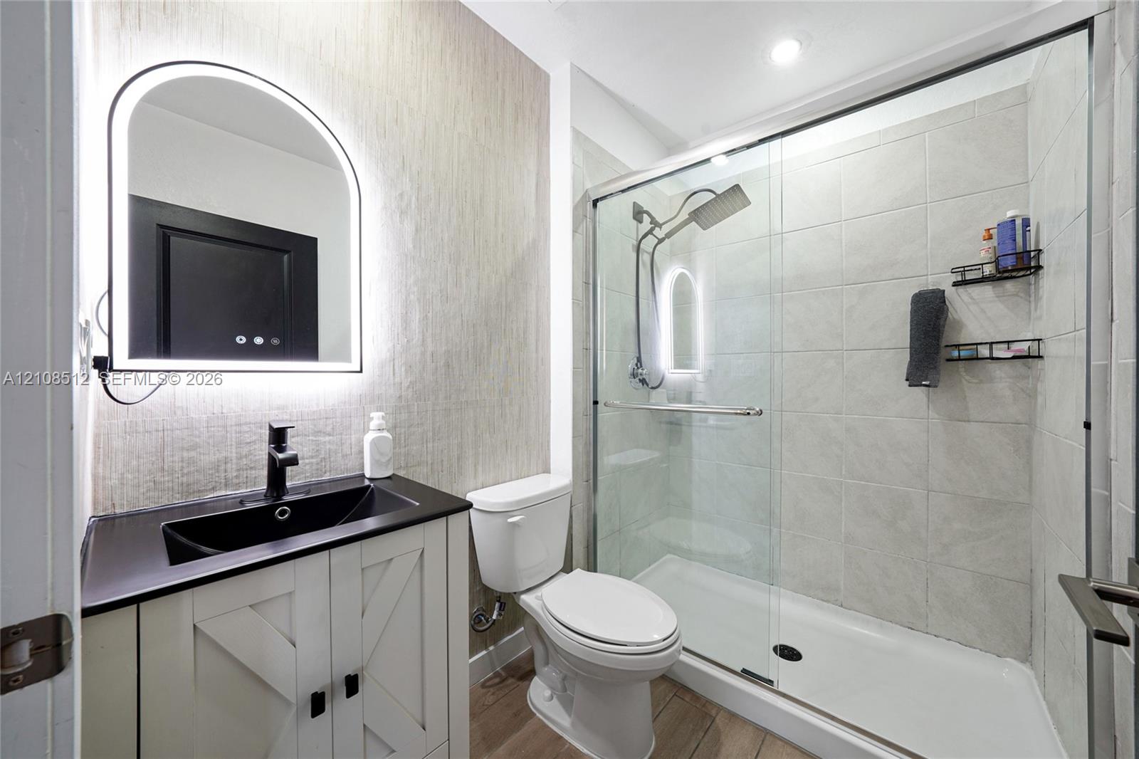 Bathroom featuring a vanity with a dark countertop and integrated sink, arch-top lighted mirror, wood-finish flooring, and a walk-in shower with a glass enclosure and tiled walls
