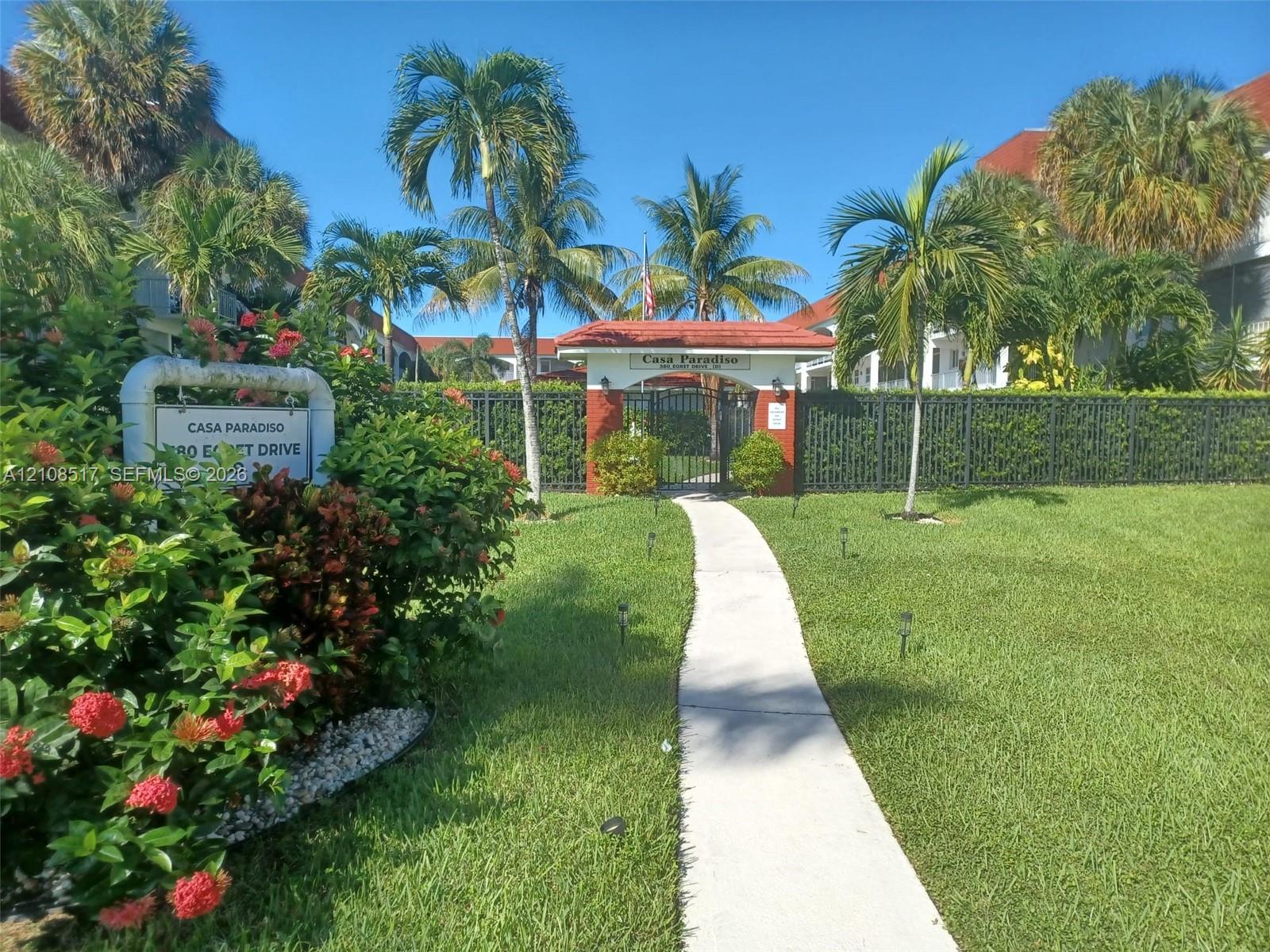 Gated community entrance featuring a red-tiled roof portico, concrete walkway, perimeter black metal fencing, and mature tropical landscaping Gated community entrance featuring a red-tiled roof portico, concrete walkway, perimeter black metal fencing, and mature tropical landscaping
