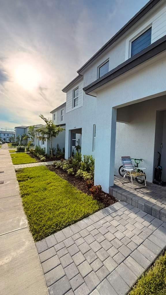 Exterior facade featuring light-toned stucco and horizontal siding, covered entryway with paver flooring, and landscaped front yard with turf and mulch beds
