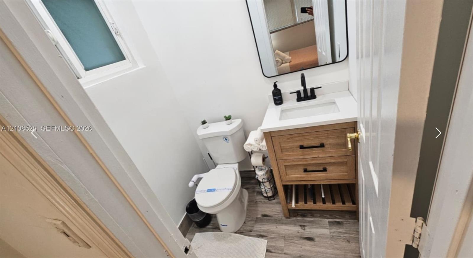 Bathroom featuring a wood-finish vanity with a white countertop and rectangular sink, matte black faucet, rectangular mirror with a black frame, and wood-finish flooring Bathroom featuring a wood-finish vanity with a white countertop and rectangular sink, matte black faucet, rectangular mirror with a black frame, and wood-finish flooring